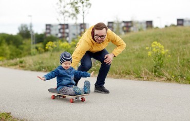 Young child sat on a skateboard being pushed along by his father.